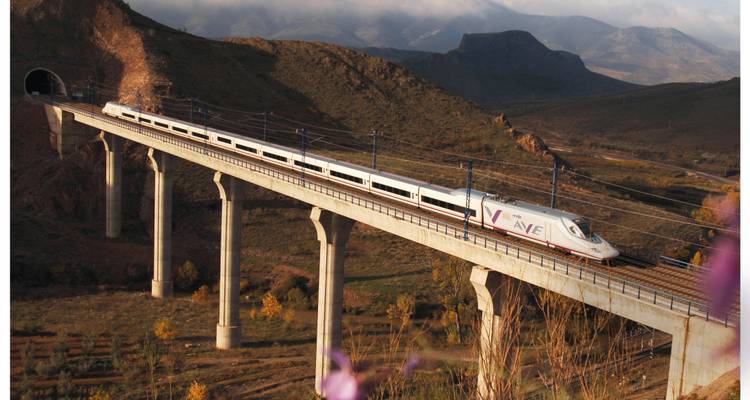 Un tren AVE de alta velocidad español cruzando un viaducto alto por un paisaje accidentado.