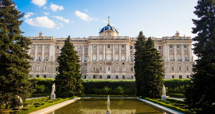 Los jardines formales y el estanque reflectante conducen hacia la majestuosa fachada del Palacio Real de Madrid bajo un cielo azul.