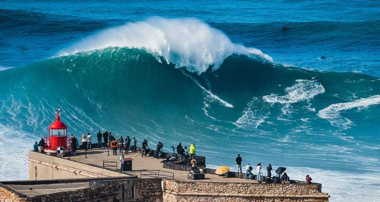 Massive Atlantikwelle stürzt neben dem roten Leuchtturmfort in Nazaré ab, während ein Surfer die Welle reitet