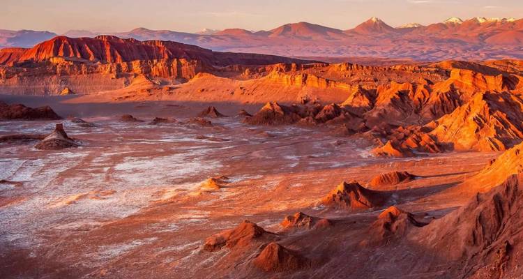 Les formations désertiques rouges et brunes étendues de la Vallée de la Lune brillant au coucher de soleil avec les pics andins lointains.