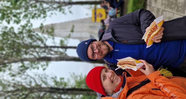 Smiling couple in beanies enjoying Icelandic hot dogs with Hallgrimskirkja rising softly in the background