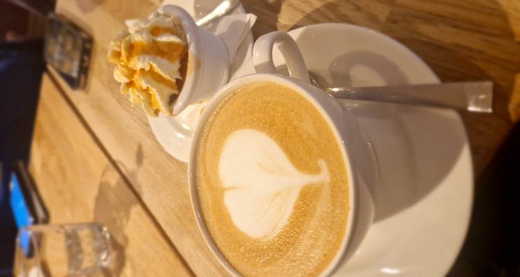 Cup of cappuccino with heart-shaped latte art beside a small dish topped with whipped cream on a wooden table