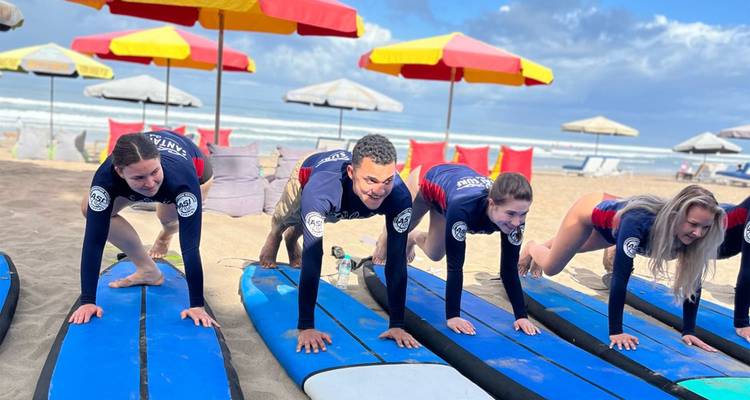 Los estudiantes de surf practican salidas en tablas azules bajo sombrillas de colores en una playa arenosa.