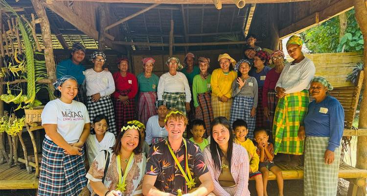 Travelers and indigenous community members pose together inside a bamboo shelter wearing colorful traditional clothing.
