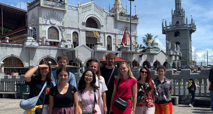 Tour group stands outside an ornate grey cathedral with towers under blue sky.