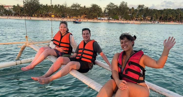 Three travelers in orange life vests relax on the outrigger net of a sailing boat near a tropical beach at sunset.