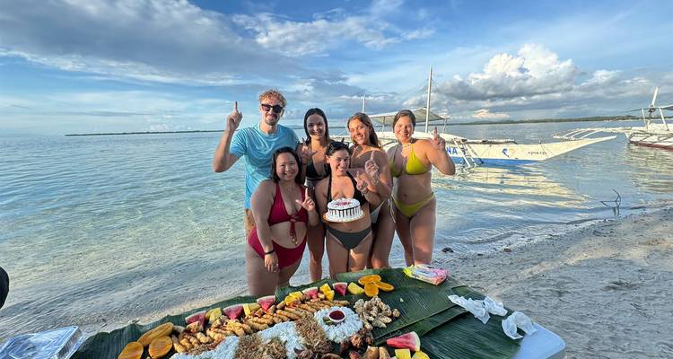 Birthday celebration on a calm beach: friends in swimwear posing with cake beside a spread of tropical fruits on banana leaves.