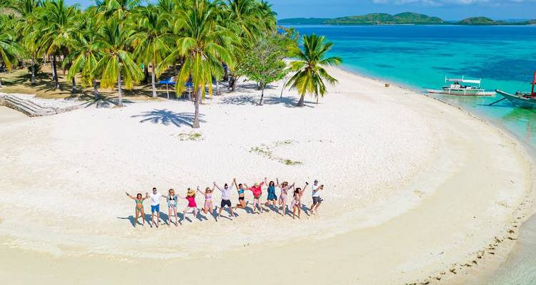 Drone shot of tiny white-sand island fringed by palms in vivid blue sea with tour group lined up in the shape of a smile.