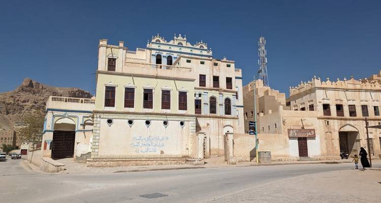 Edificios históricos decolorados por el sol se alinean en una calle tranquila bajo un cielo azul intenso; una mujer solitaria pasa caminando.