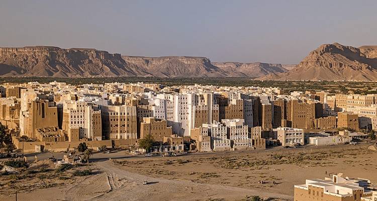 Vista aérea de los imponentes rascacielos de ladrillo de barro de Shibam ubicados en una llanura desértica con acantilados detrás.