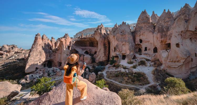 Female traveler with hat stands on a rock ledge admiring cave dwellings and spires of Cappadocia
