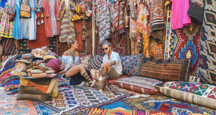 Two travelers sitting among stacks of vivid Turkish carpets and pillows inside a colorful bazaar stall