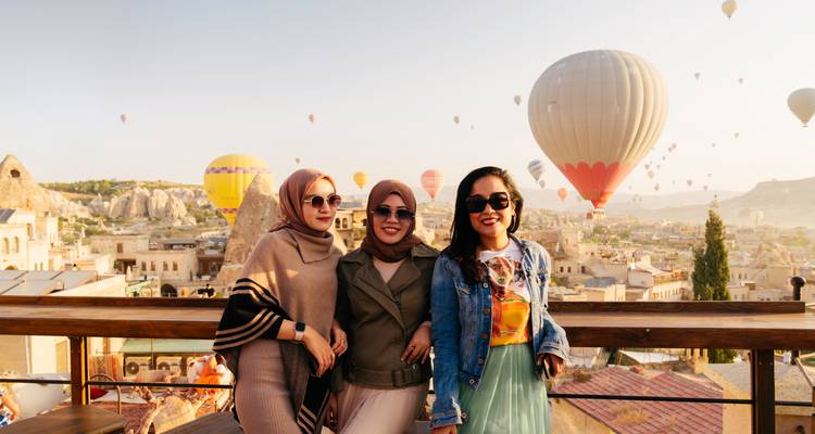 Three friends pose on a rooftop terrace while colorful balloons rise over the unique landscape of Cappadocia.