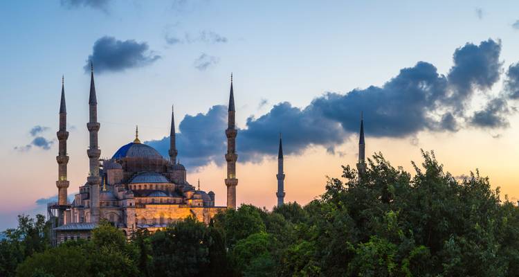 Blue Mosque at dusk, softly illuminated beneath dramatic clouds and framed by trees