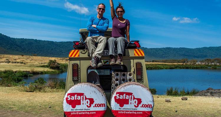 Two travelers sitting on the rear of a safari vehicle beside a scenic crater lake with branded spare-tire covers.