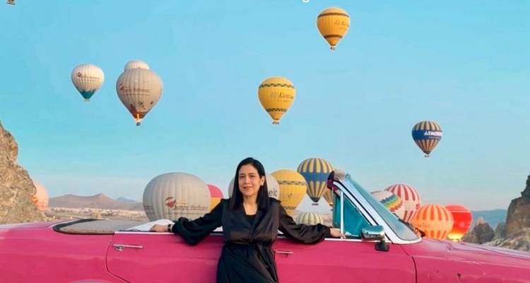 Mujer recostada contra un coche vintage rosa mientras múltiples globos aerostáticos coloridos flotan en el cielo sobre Capadocia
