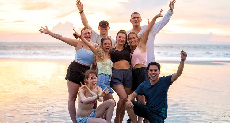 Vrolijke groep jonge reizigers poseert op een tropisch strand bij kleurrijke zonsondergang met zachte golven achter hen.