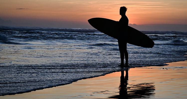 Silhouette eines Surfers, der ein Brett hält und knöcheltief im reflektierenden Wasser während eines flammenden Sonnenuntergangs steht.