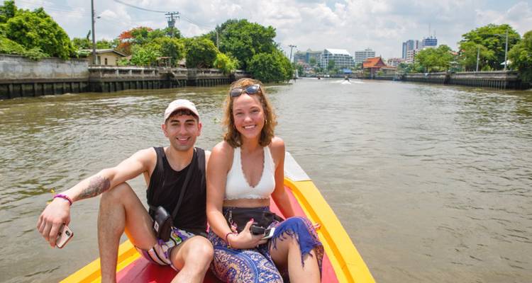 Twee mensen die op een boot zitten in een rivier met stadsgebouwen op de achtergrond.
