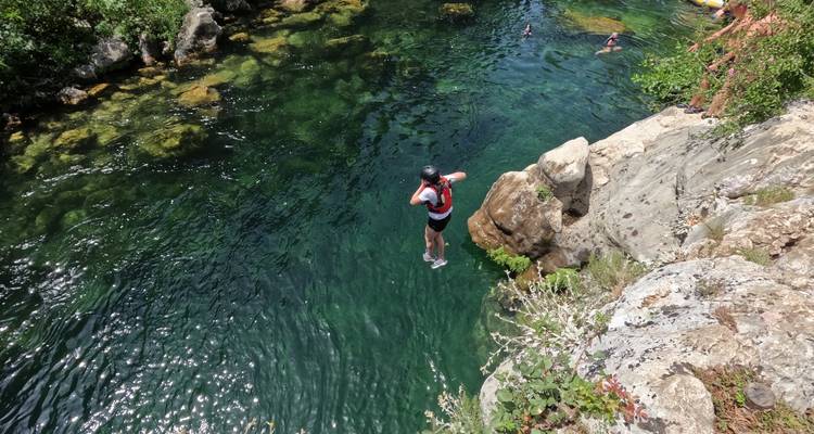 Une personne portant un gilet de sauvetage et un casque saute d'un rocher dans une gorge de rivière émeraude cristalline entourée de falaises rocheuses et d'arbustes.