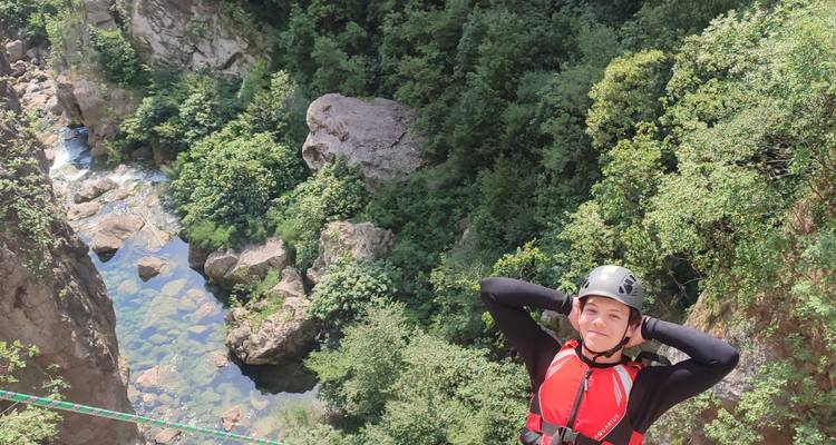 Aventurier souriant en casque et combinaison de plongée se tenant près du bord d'une falaise surplombant une gorge boisée avec un ruisseau limpide en contrebas.