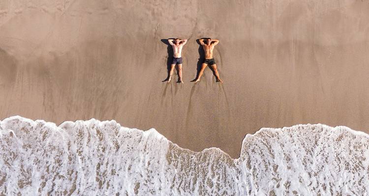 Vista aérea con dron de dos personas tomando el sol acostadas en una playa dorada justo más allá de la orilla espumosa.