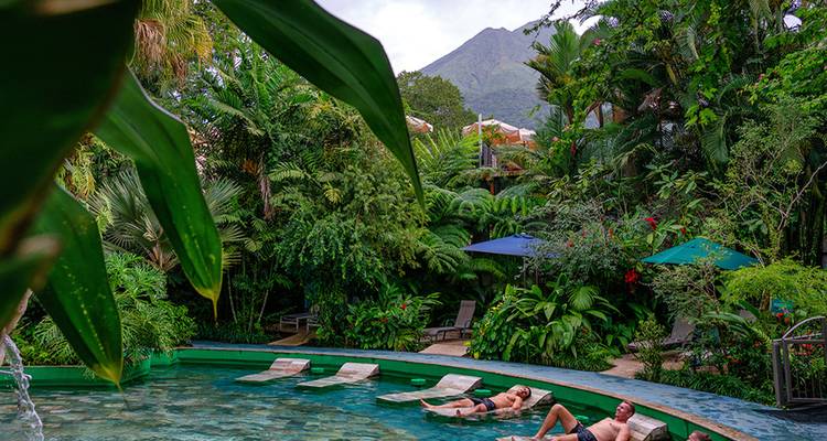 Pareja se relaja en asientos de descanso en una piscina tropical ubicada en exuberantes jardines con un volcán elevándose al fondo.