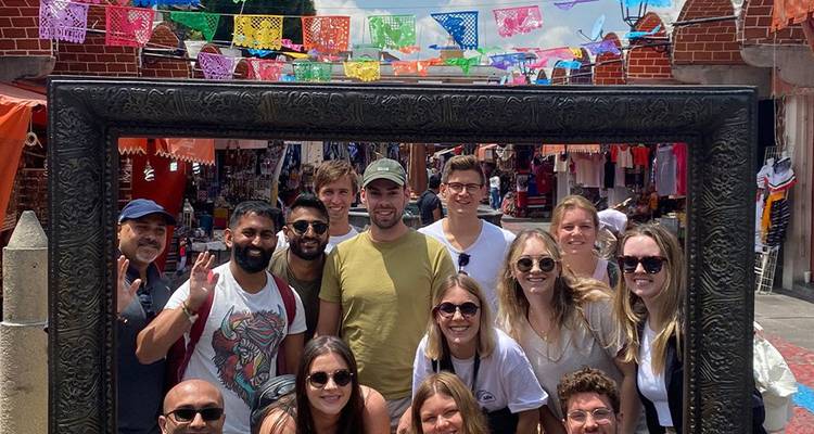 Groep reizigers die poseren in een groot decoratief fotolijstje op een kleurrijke straatmarkt versierd met papel picado.