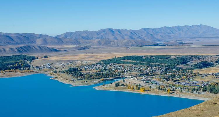 Le Lac Bleu Tekapo s'étendant à côté d'une petite municipalité avec des plaines arides et des chaînes de montagnes au-delà