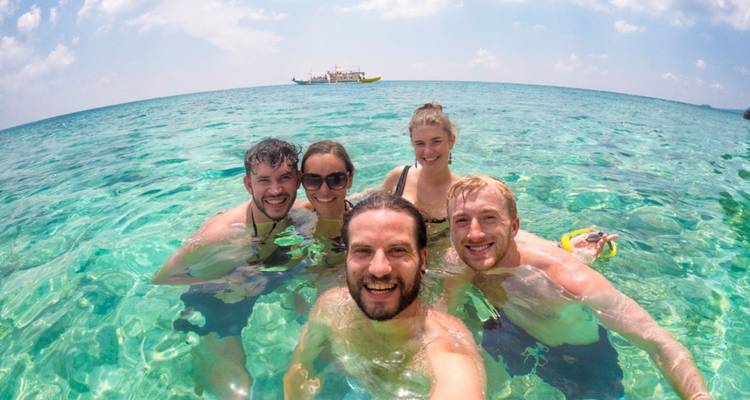 Cinco amigos jóvenes se toman una selfie alegre mientras nadan en agua de mar turquesa cristalina con un bote de tour de fondo.