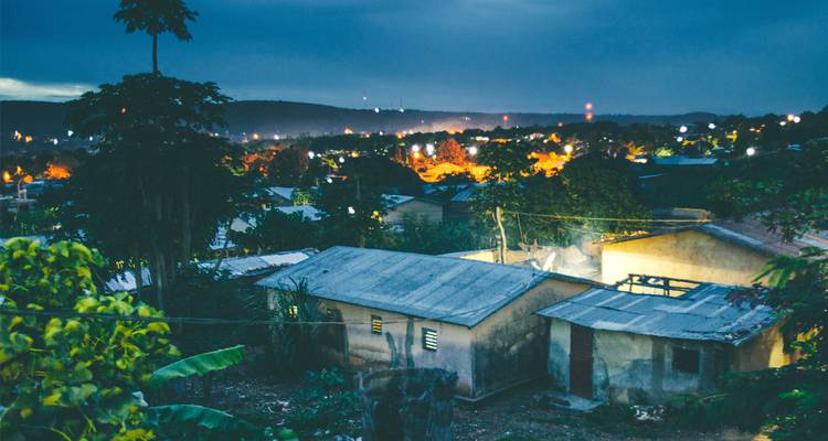 Vista nocturna de un asentamiento rural con casas de techo de hojalata y luces lejanas del pueblo bajo un cielo azul oscuro.