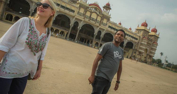 Twee reizigers glimlachen op het open terrein van het Mysore Palace op een zonnige dag.