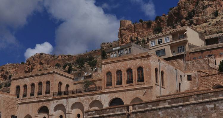Las casas de piedra arenisca y edificios históricos suben por una ladera rocosa bajo un cielo azul brillante y nubes esponjosas.