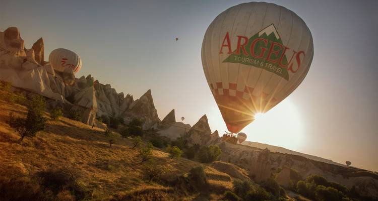 Un globo aerostático flotando sobre las extrañas torres de roca de Capadocia al amanecer con la cálida luz del sol brillando detrás del globo.