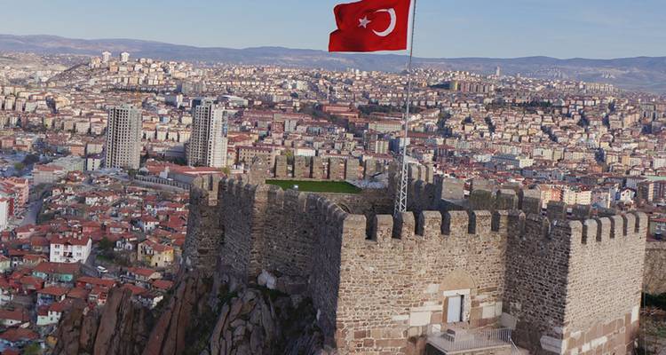 Muros de piedra del Castillo de Ankara con una gran bandera turca ondeando, dominando la extensa ciudad moderna bajo cielos despejados.