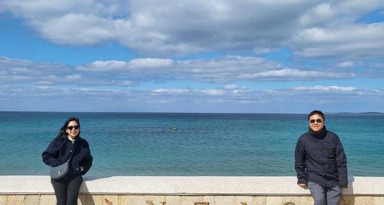 Pareja posando en un paseo marítimo con agua turquesa y cielo parcialmente nublado detrás de ellos