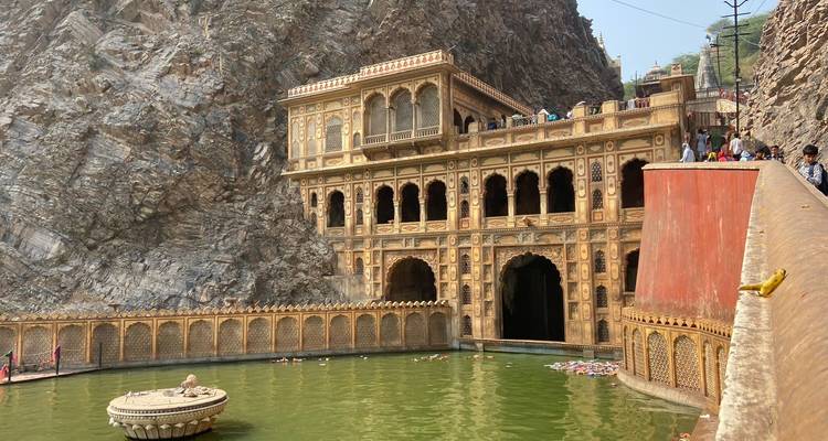 Historic Galtaji temple complex built into sheer rock, with green sacred pool and visitors around.