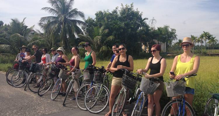 Une file de voyageurs se tient debout à côté de bicyclettes sur une route rurale bordée de palmiers et de rizières.