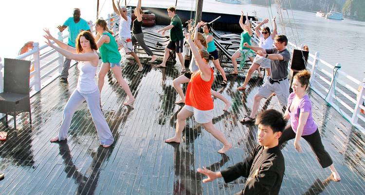 Les voyageurs pratiquent le tai-chi du matin sur le pont d'un bateau de croisière en bois avec les paysages karstiques de la baie d'Halong en arrière-plan.