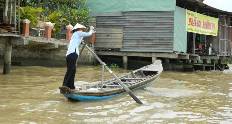 Un rameur solitaire coiffé d'un chapeau conique propulse une embarcation en bois élancée devant des bâtiments riverains sur pilotis dans les eaux troubles du Mékong.