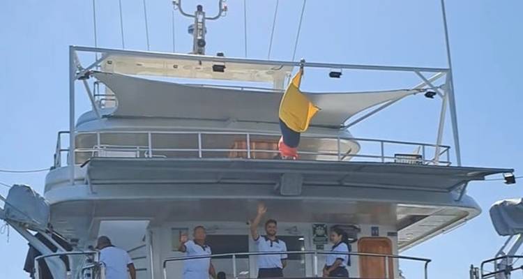Crew members waving from the deck of a yacht beneath a national flag on a clear day
