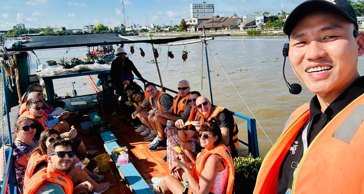 Ein lächelnder Reiseführer macht ein Selfie mit Reisenden in orangefarbenen Rettungswesten auf einem Boot im Mekong-Delta.