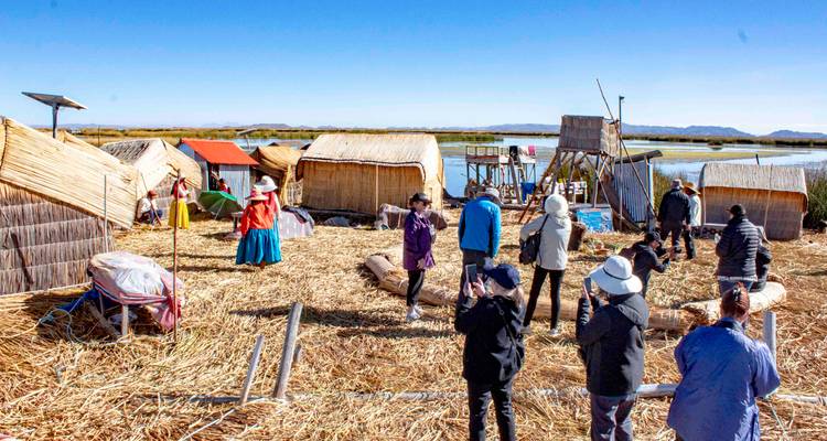 Tourists and locals standing on a reed-covered floating island with thatched huts on Lake Titicaca under a bright blue sky.