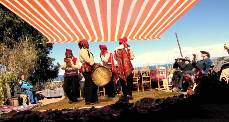 Traditional musicians perform under a striped canopy while tourists watch on the shore of Lake Titicaca.