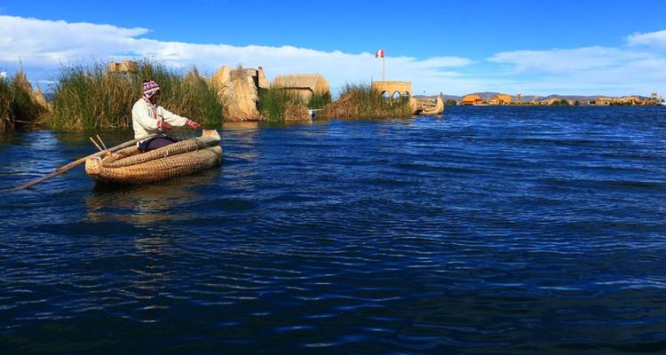 Man paddles a traditional reed canoe across the deep blue waters of Lake Titicaca beneath a clear sky.