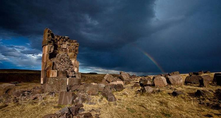 Lone ancient stone tower stands amid rocky grassland under stormy clouds with a rainbow arching across the sky.