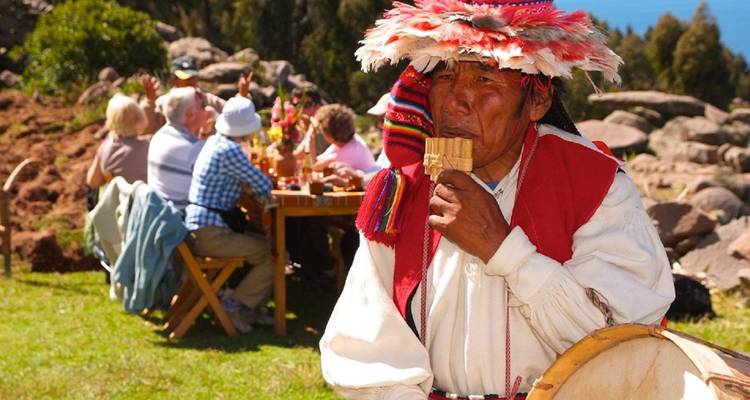 Man in colorful headdress plays panpipes in the foreground while visitors enjoy an outdoor meal in the background.