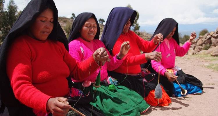 Four women in bright traditional attire kneel together, spinning yarn with hand spindles against a rural backdrop.