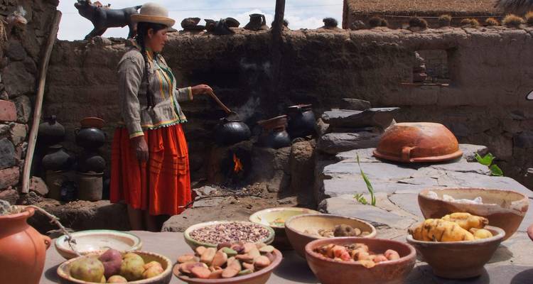 Woman in traditional dress cooks over an open fire in an adobe kitchen surrounded by bowls of local produce.