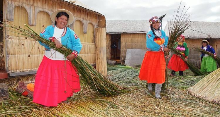 Smiling women on a reed island carry bundles of fresh green reeds beside thatched houses.